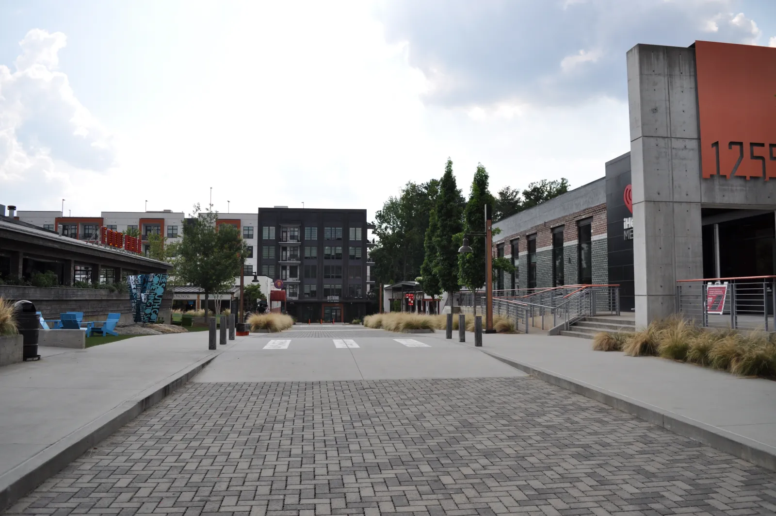 Wide pedestrian plaza with modern commercial buildings, trees, and textured pavement under partly cloudy skies.