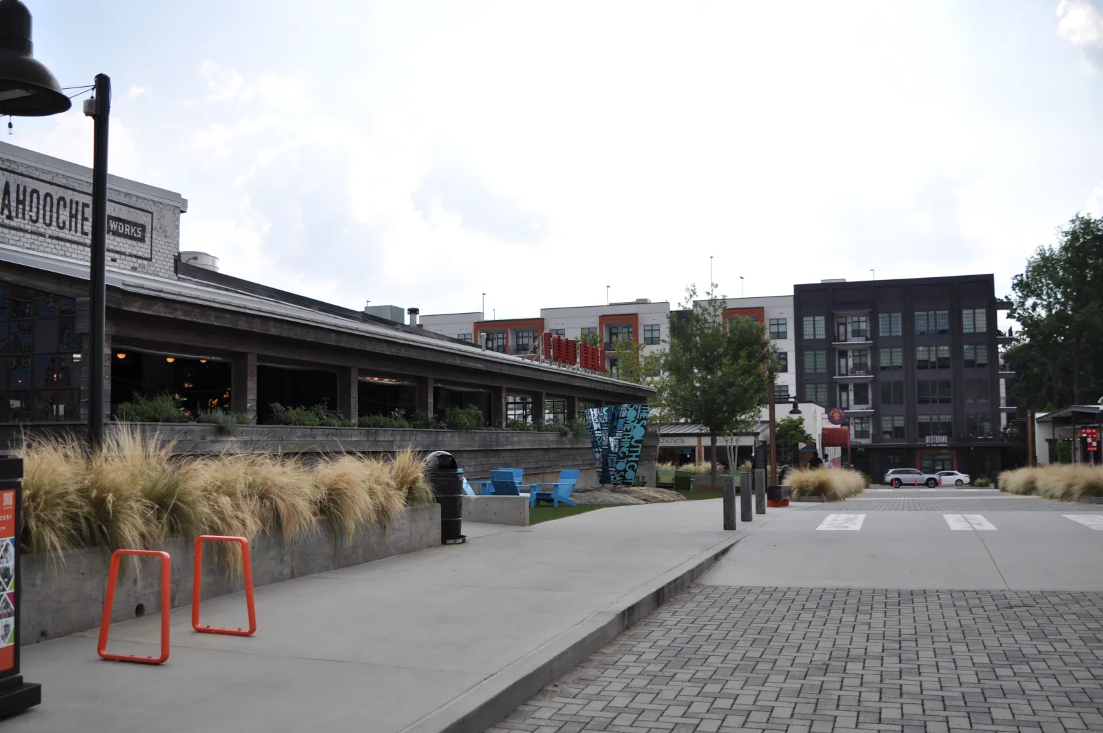 Outdoor urban scene with modern buildings, a restaurant patio, ornamental grasses, and pedestrian pathways under a partly cloudy sky.