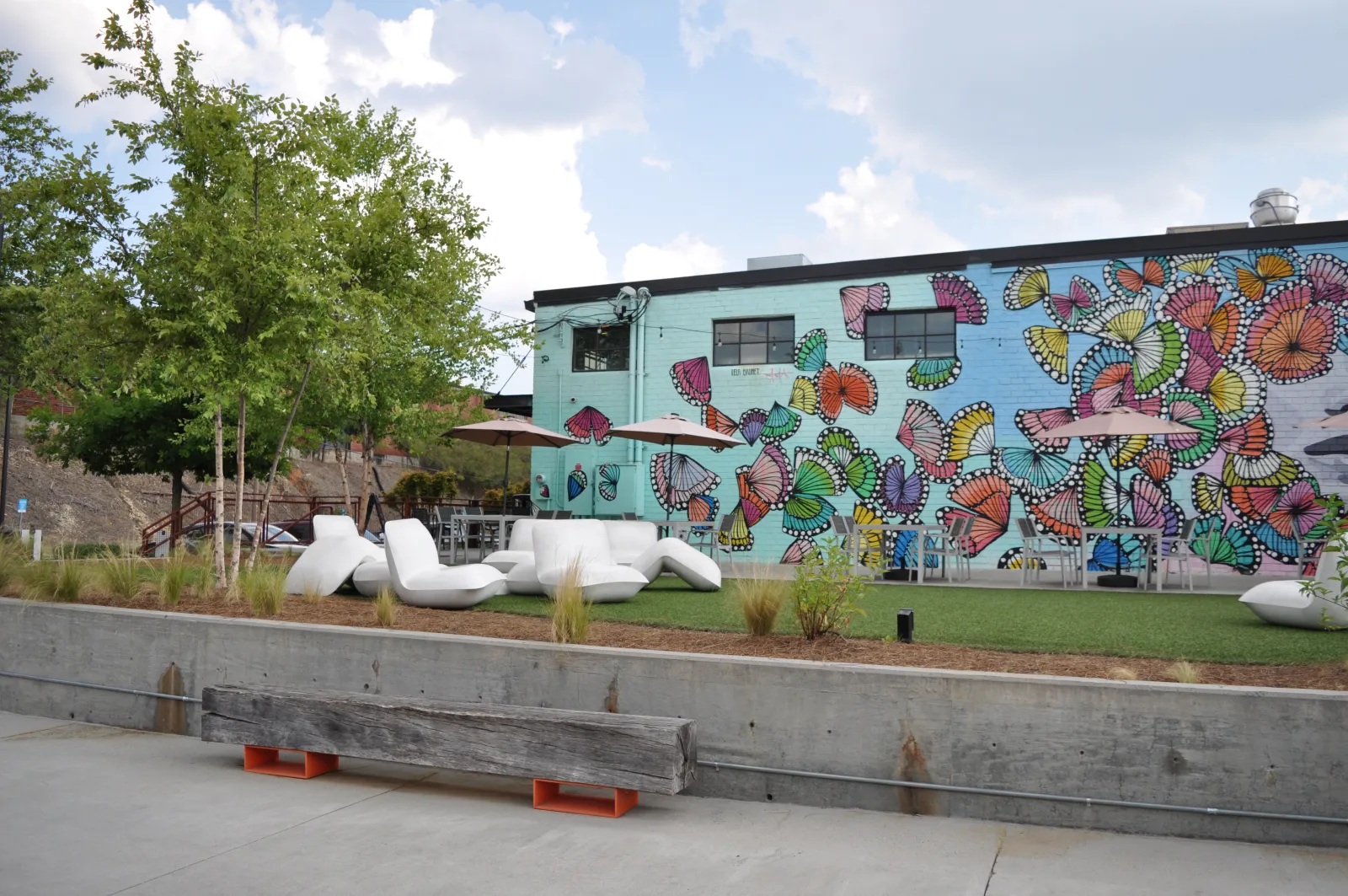 Outdoor seating area with modern white furniture and umbrellas in front of a colorful butterfly mural on a building wall.