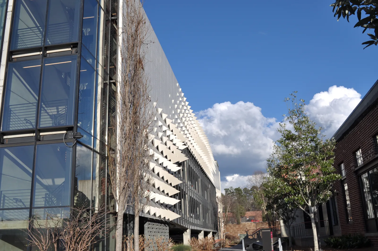 Modern building with glass panels and geometric facade under a blue sky with clouds and surrounding trees.