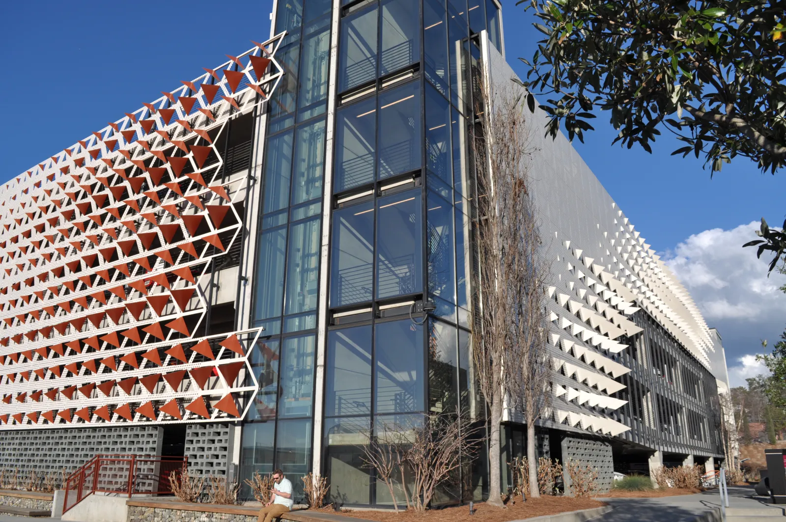 Modern building with geometric sunshade panels on facade and glass elevator tower under clear blue sky.