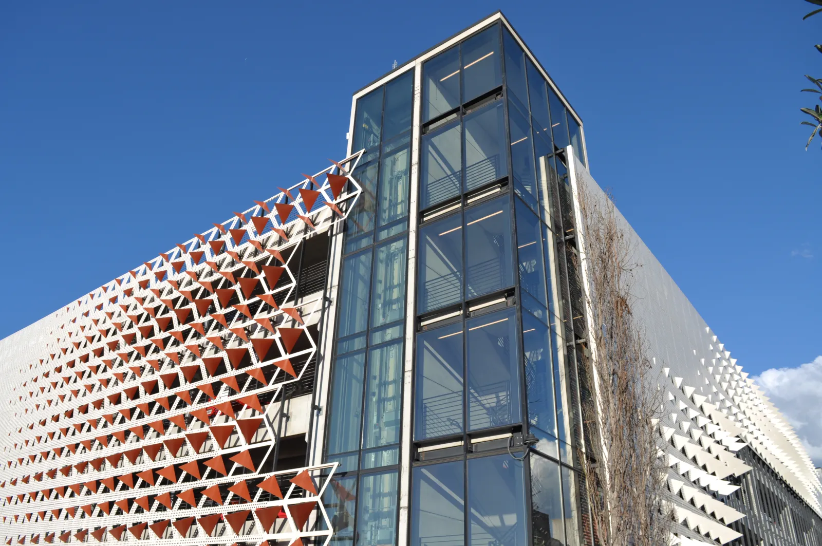 Modern building facade with geometric sunshades and glass elevator tower under clear blue sky