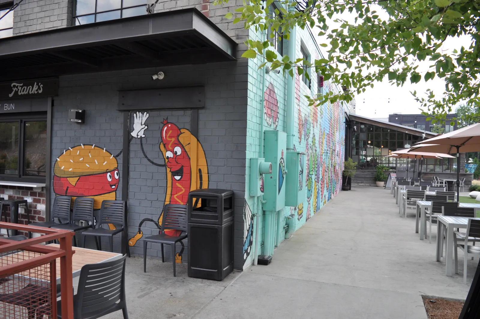 Outdoor seating area with mural of cartoon burger and hot dog high-fiving on brick wall at restaurant.