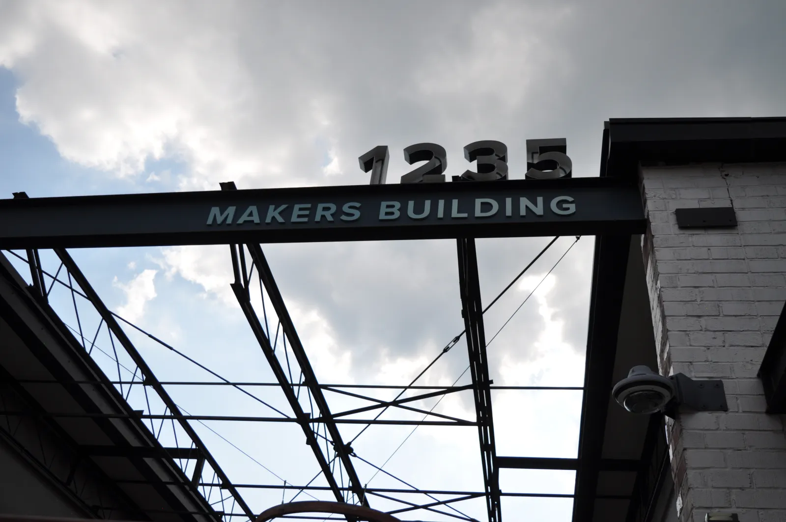 Structural metal beam with Makers Building sign and 1235 number against cloudy sky background.