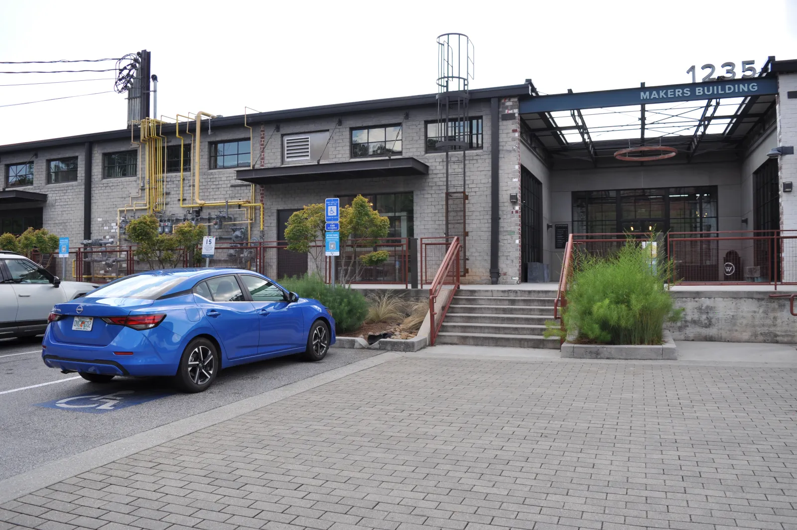 Blue sedan parked in front of a gray industrial Makers Building with stairs and greenery on a cloudy day