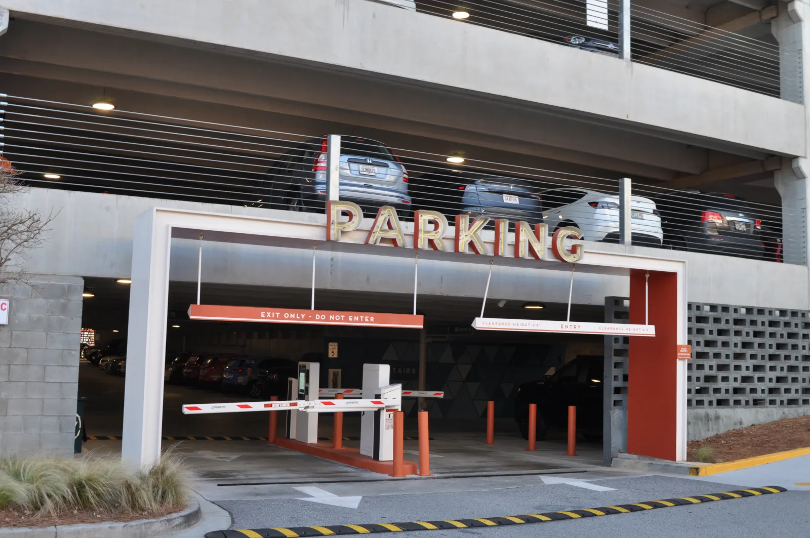 Entrance to a multi-level parking garage with barrier gates and parked cars above in daylight.