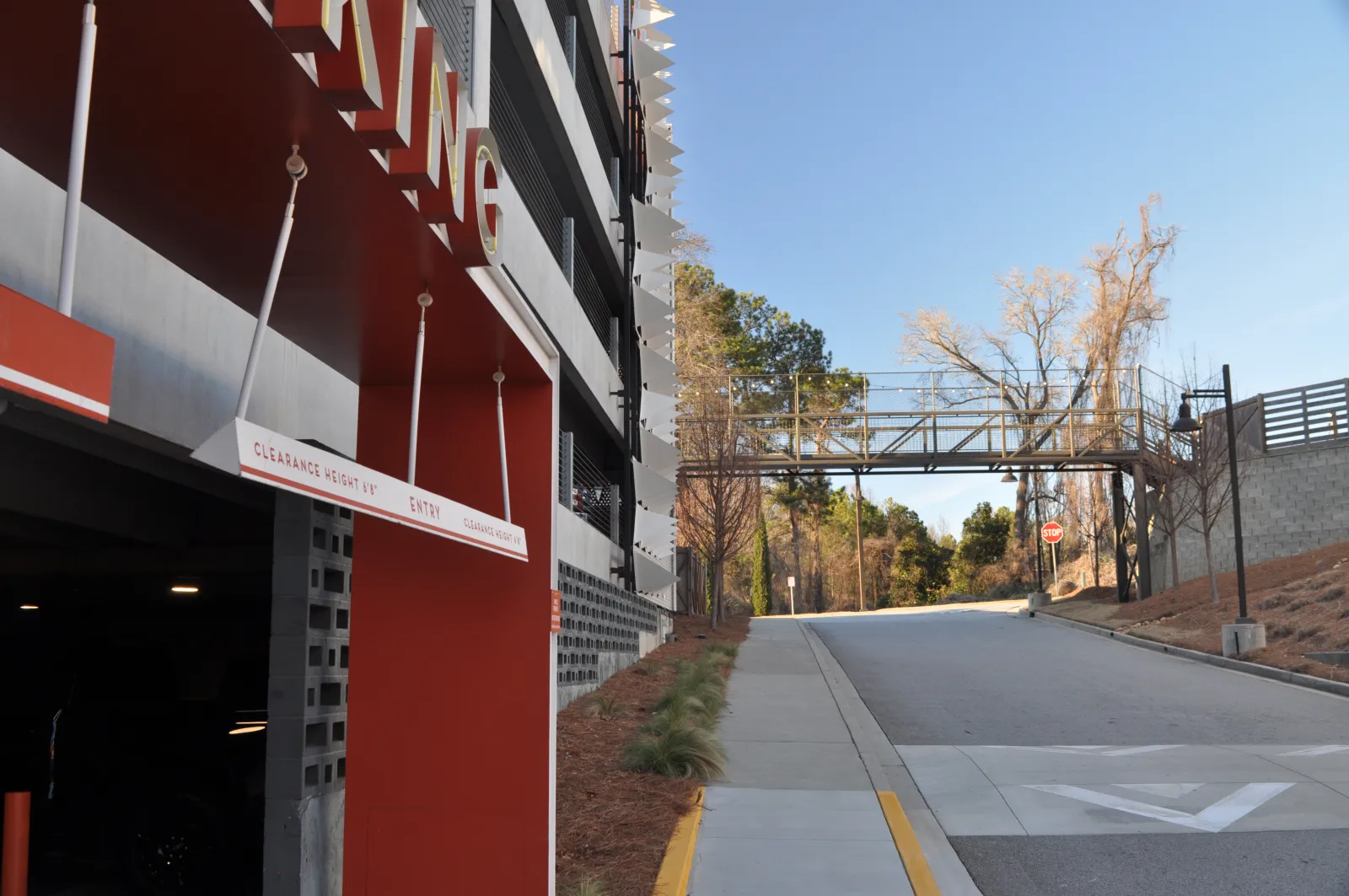 Parking garage entrance with clearance height sign next to a road leading to a pedestrian bridge over the street.
