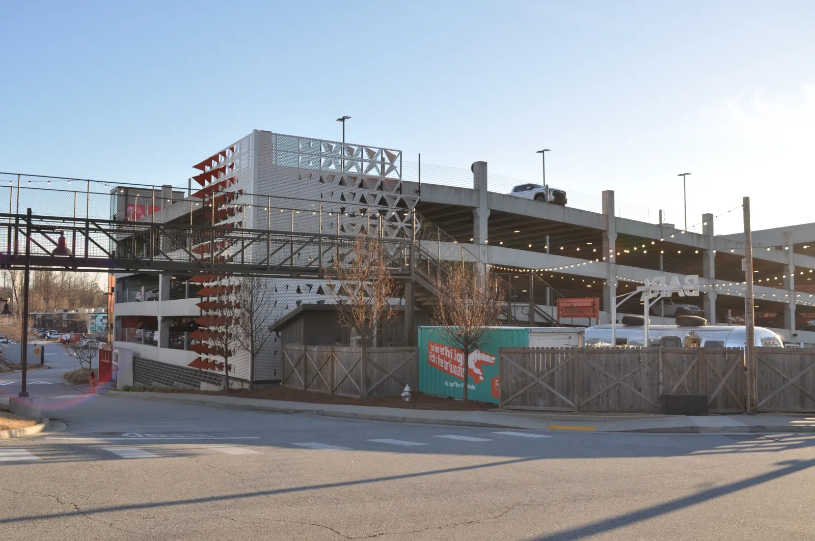 Outdoor multi-level parking structure with decorative panels, hanging lights, and fenced area at sunset.