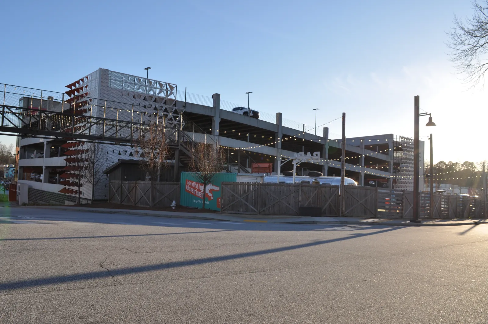 Multi-level parking garage with string lights and wooden fence under a clear blue sky during daytime.