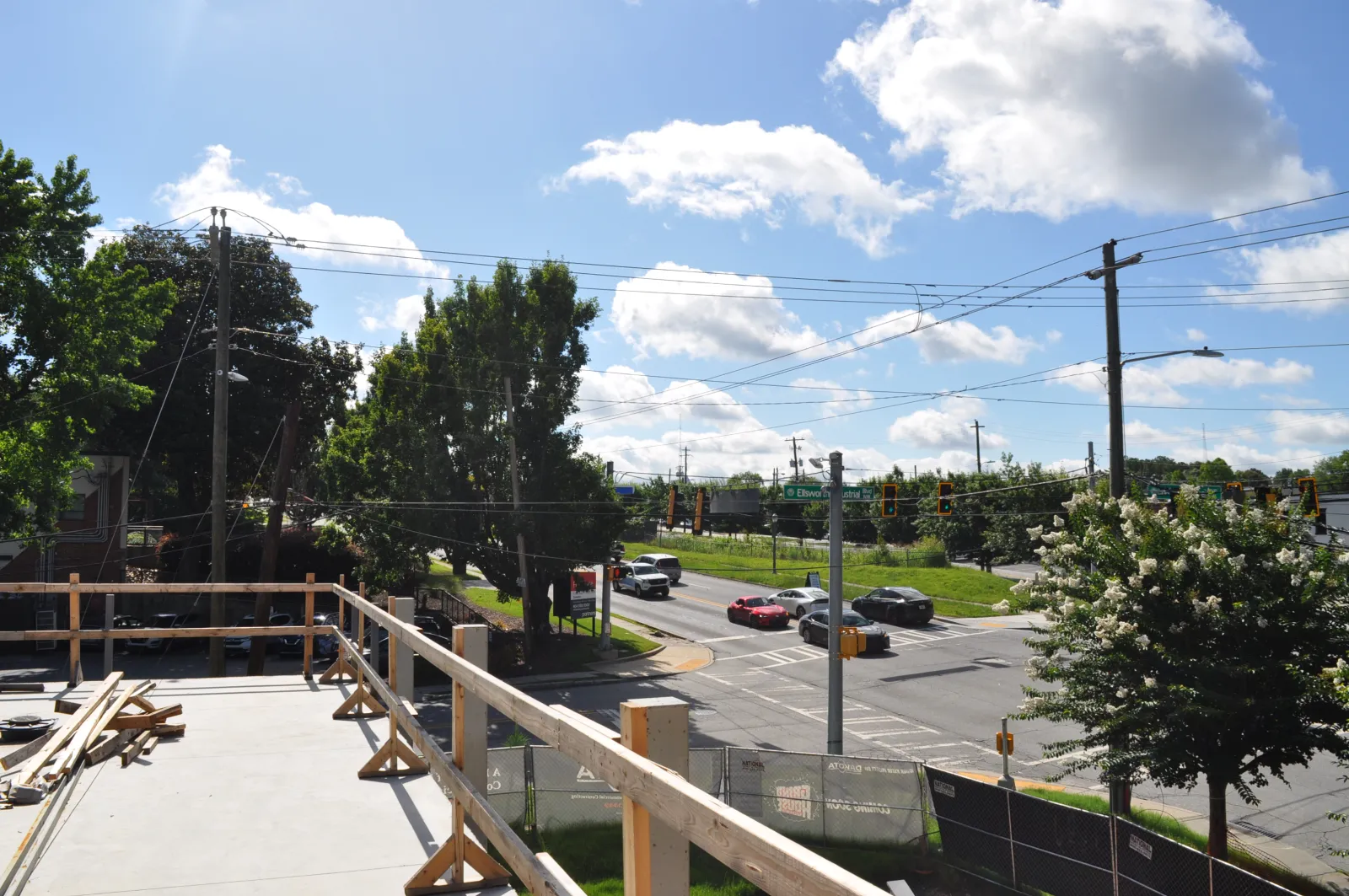 View of a sunny suburban intersection with cars, trees, and construction site railing in the foreground.