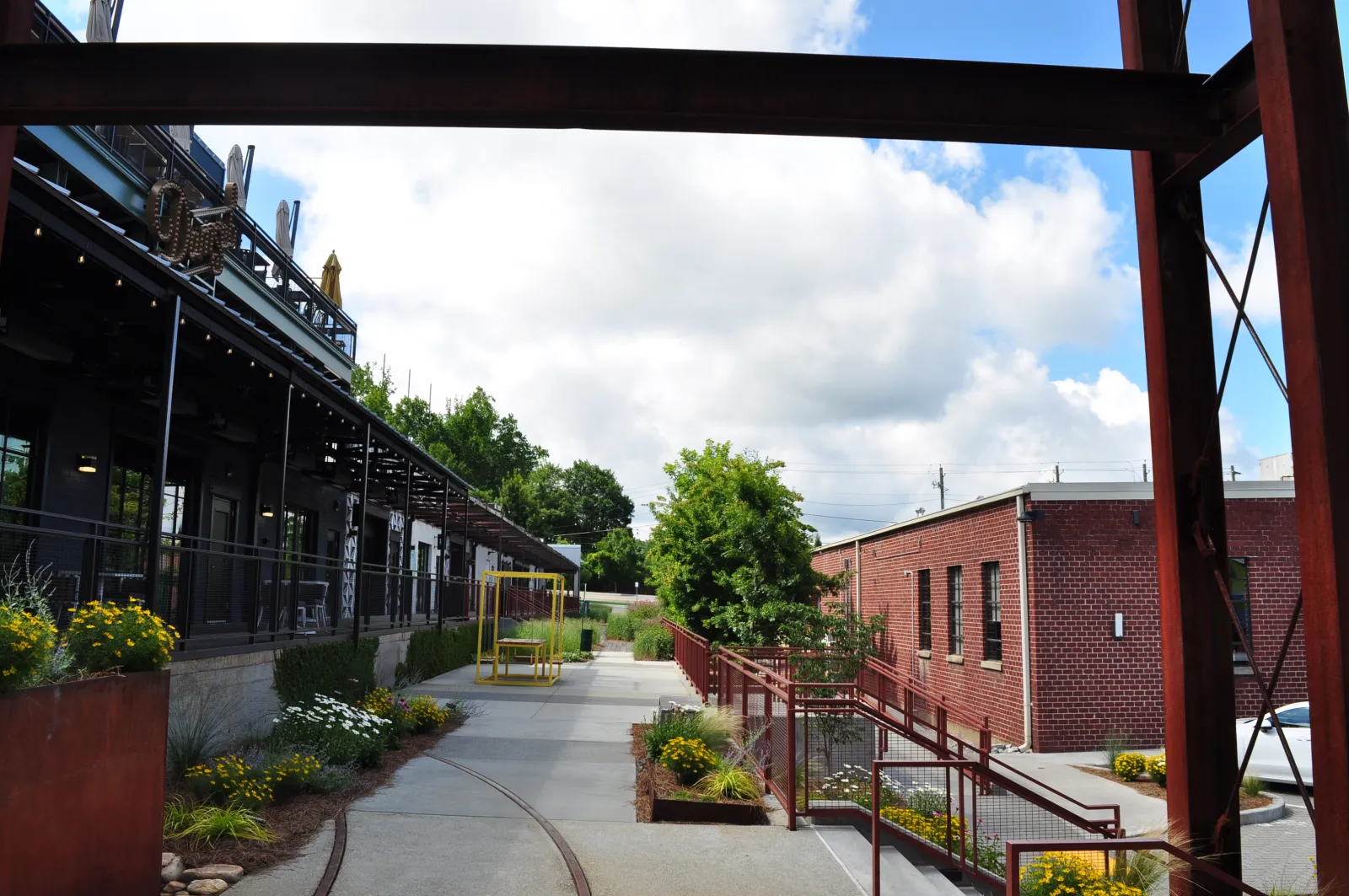 Outdoor walkway between modern commercial buildings with greenery, flowers, and partly cloudy sky overhead