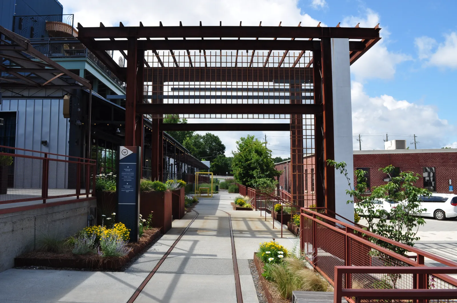 Modern outdoor walkway with metal pergola, planters, and urban buildings under a blue sky with clouds.
