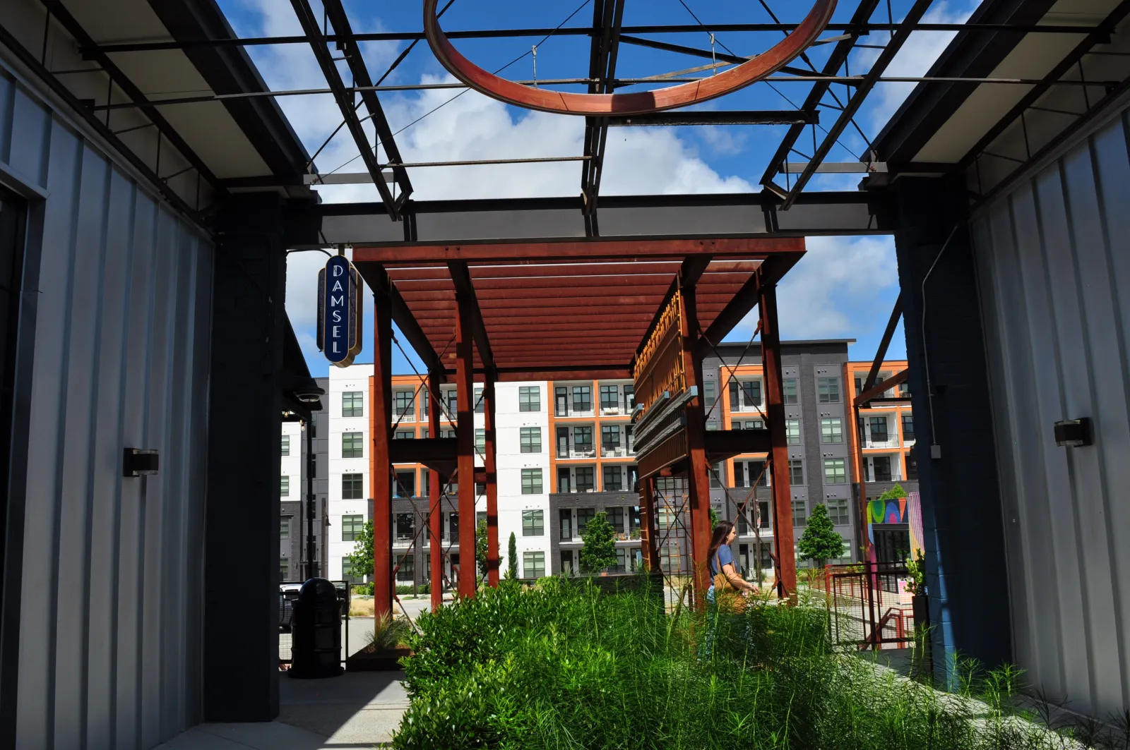 Outdoor modern urban area with metal structures, greenery, and a person walking near apartment buildings on a sunny day