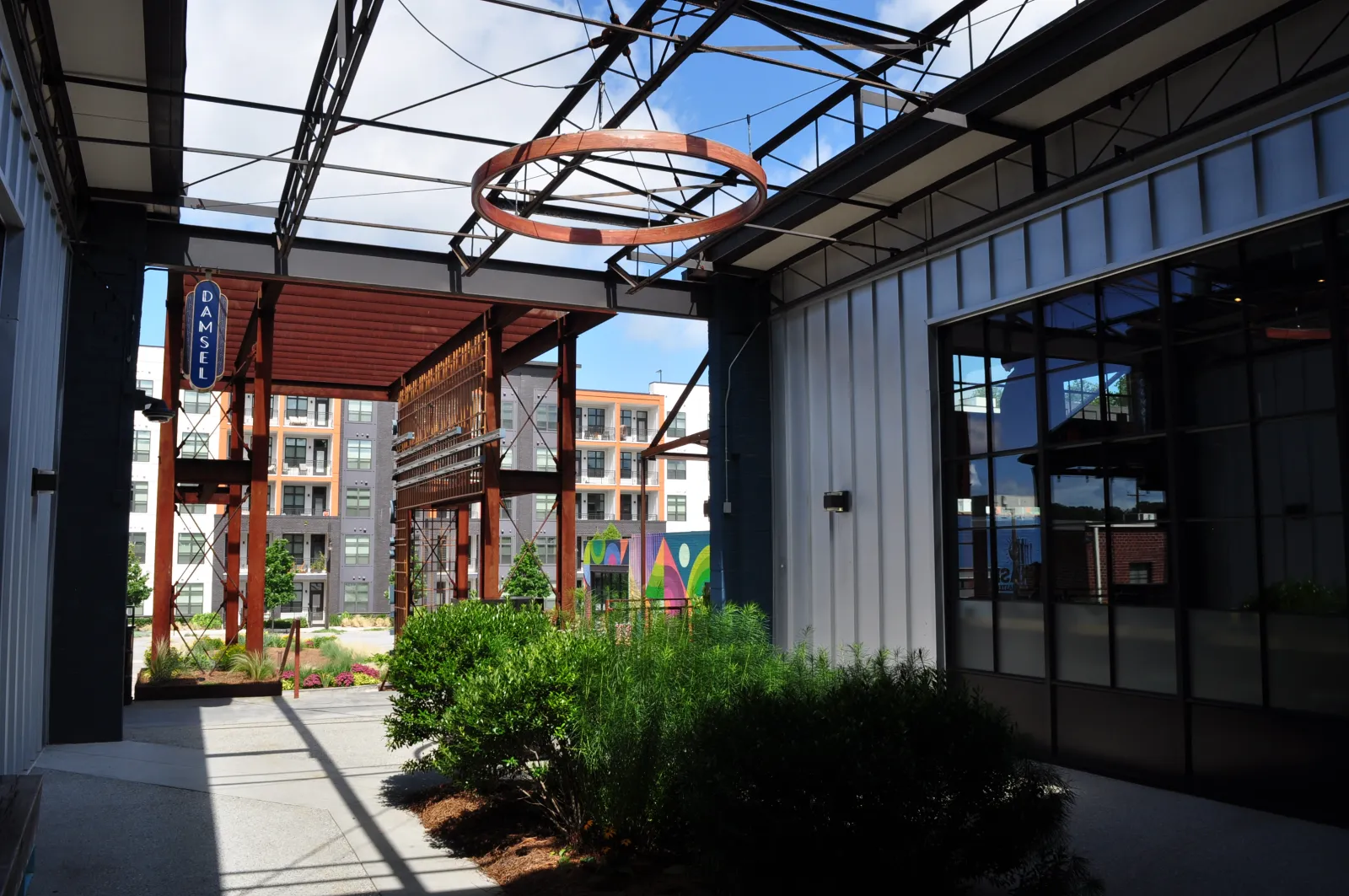 Modern industrial-style outdoor corridor with greenery, metal framework, glass windows, and apartment buildings in background