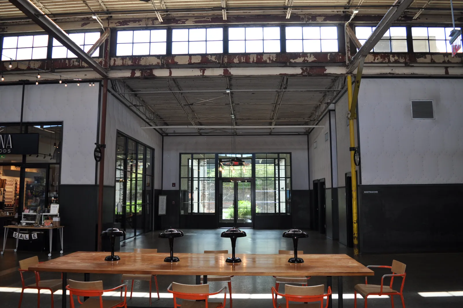 Spacious industrial-style room with large windows, high ceiling, long wooden table, and orange chairs under natural light.