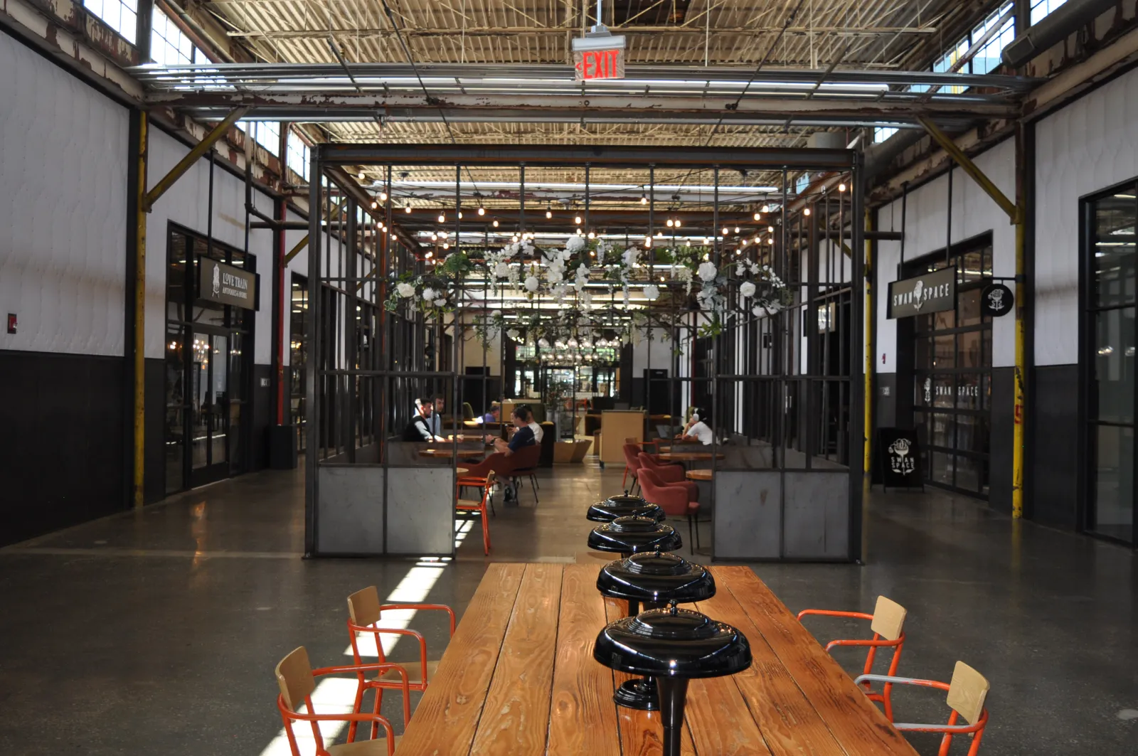 Open industrial-style coworking space with wooden tables, orange chairs, hanging plants, and exposed ceiling lighting.
