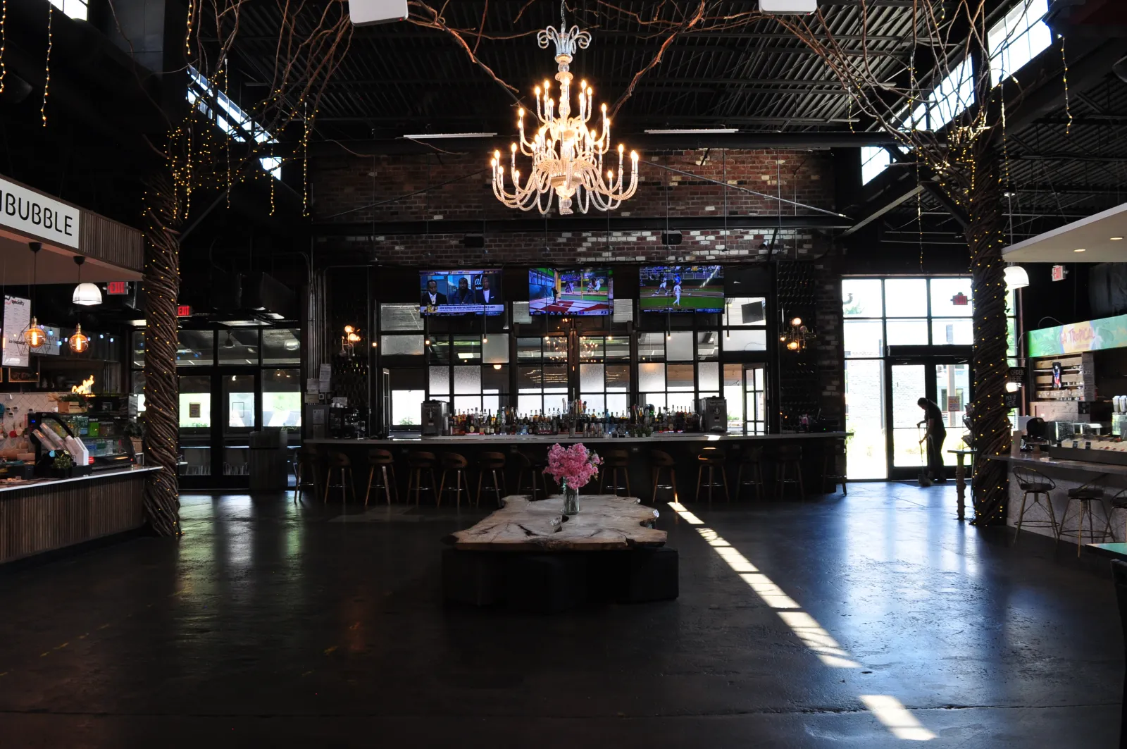 Spacious modern bar with wooden accents, chandelier, multiple TVs, and pink flower centerpiece on large wooden table.