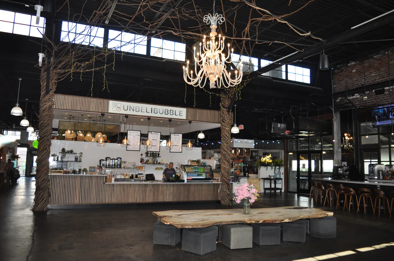 Modern cafe interior featuring a wooden bench table, chandelier, and UnbeliBubble counter under tall windows.