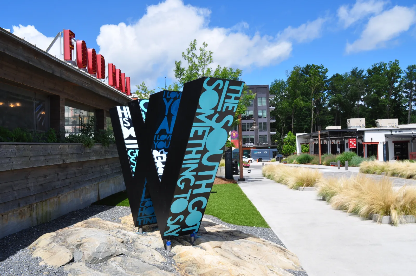 Large decorative 'W' sculpture with text in an outdoor urban park area under blue sky.