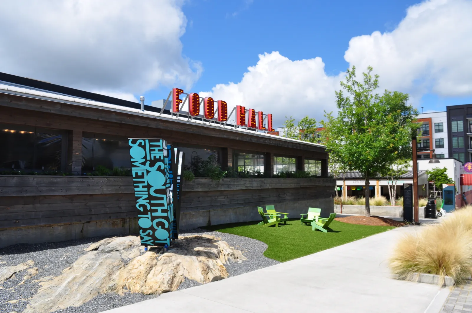 Outdoor seating area with green chairs and a wooden food hall under a blue sky with clouds