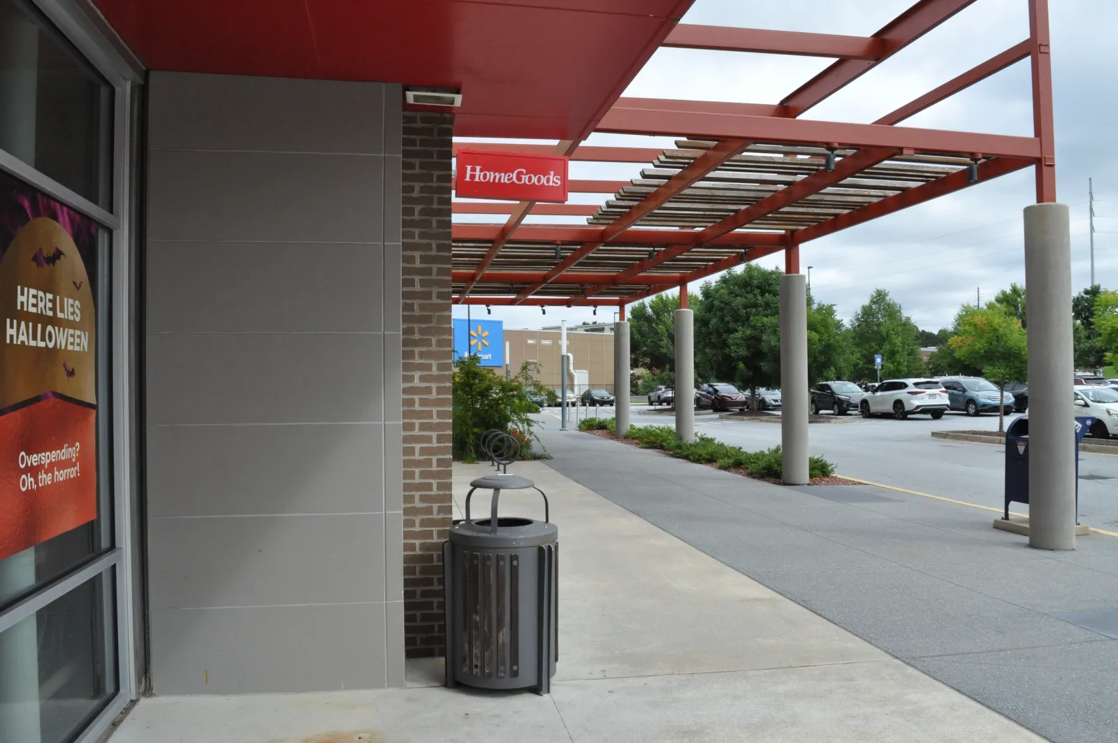 Outdoor shopping area with HomeGoods sign, parking lot, trash can, and Halloween sale advertisement on storefront window.