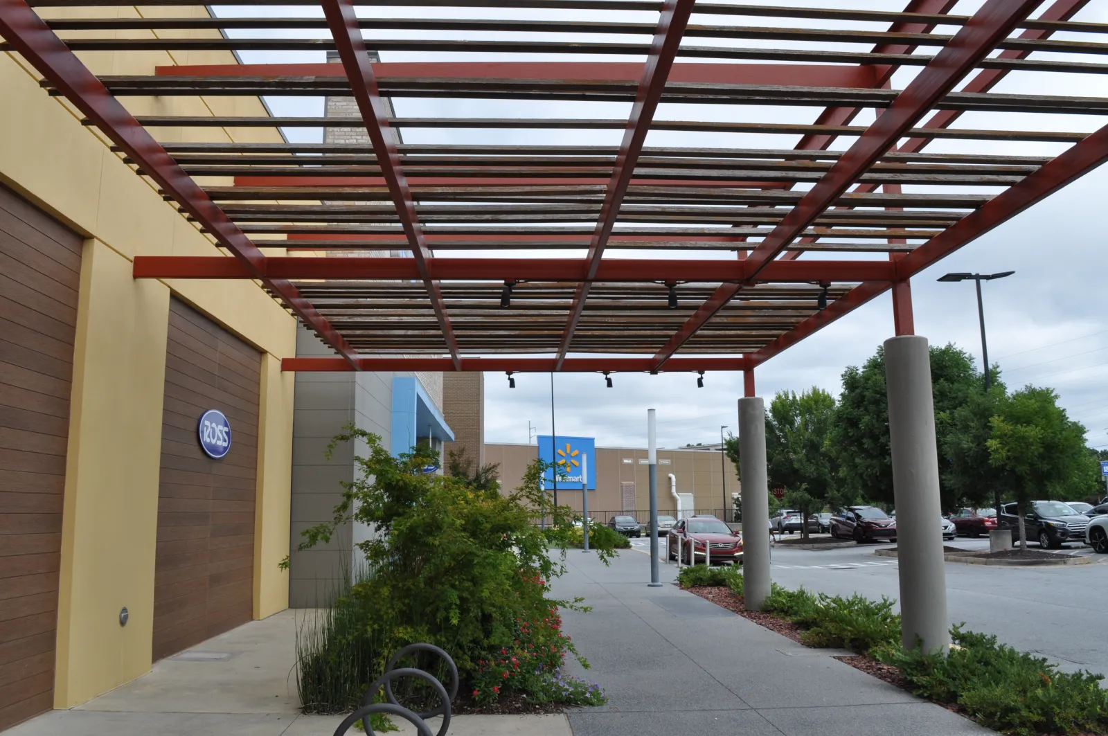 Outdoor shopping area with Ross and Walmart stores, covered walkway, plants, and parked cars on a cloudy day