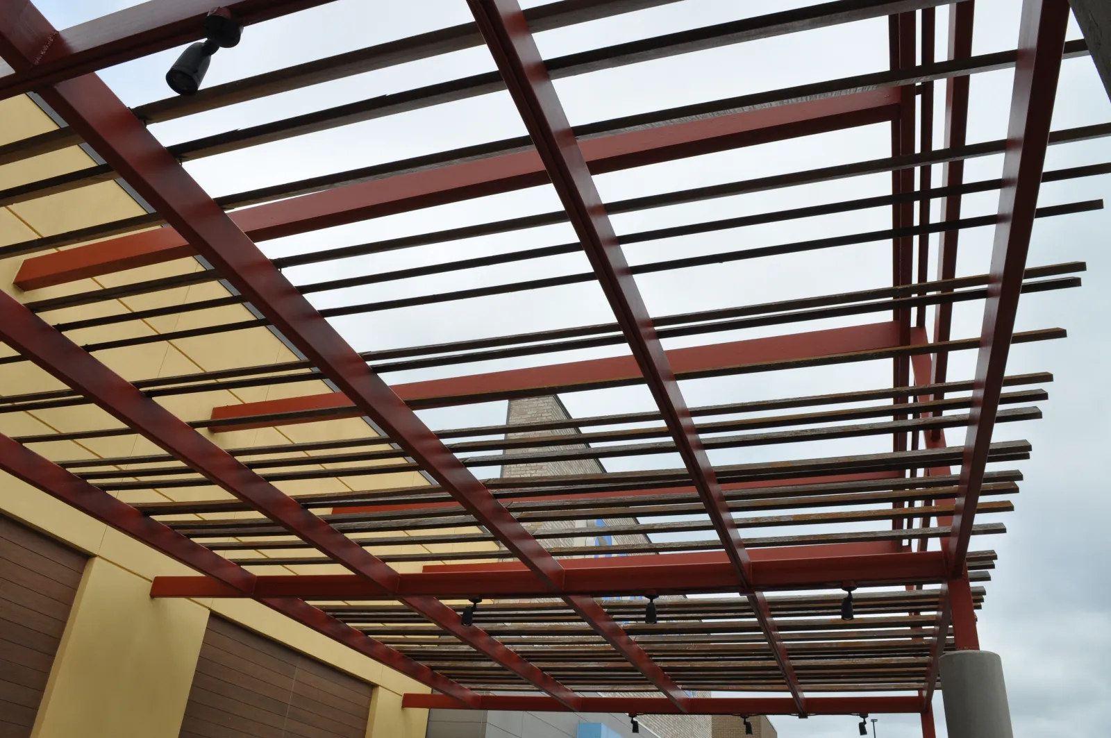 Modern pergola with wooden slats and red beams over a building exterior under cloudy sky.