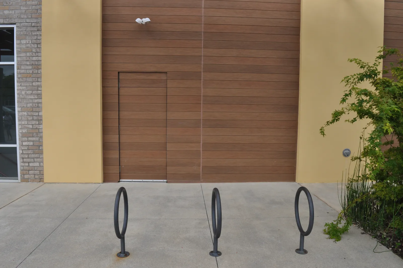 Empty bike racks in front of a modern wooden and yellow building wall with a concealed door and greenery.