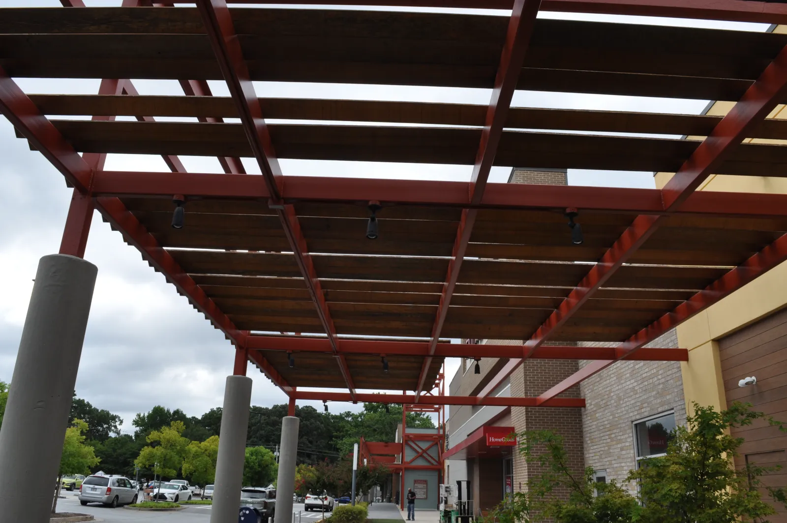Outdoor walkway with red steel pergola and wooden slats next to a brick building under cloudy sky.