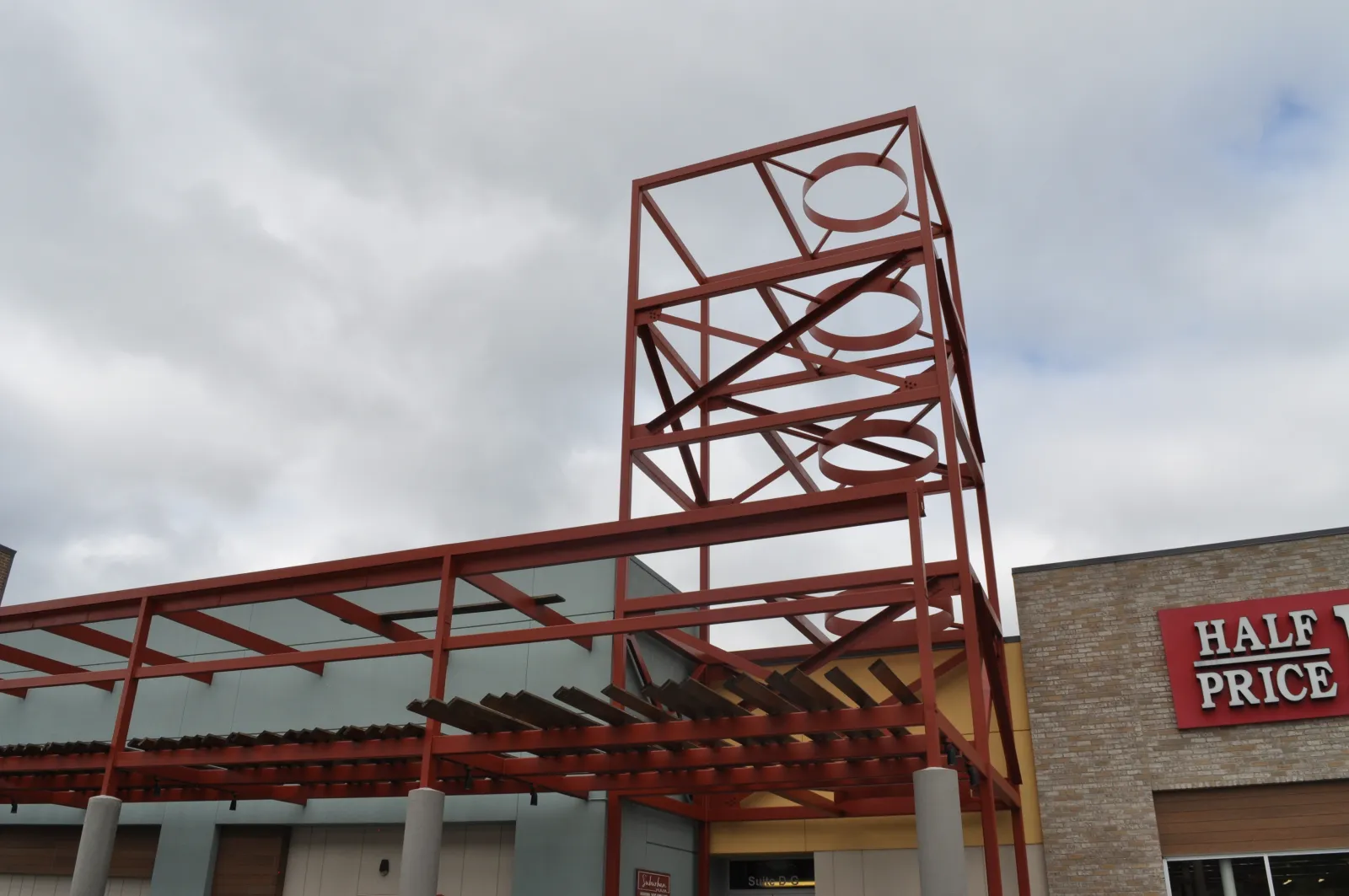Red metal framework structure with circular cutouts above a building entrance under cloudy sky.