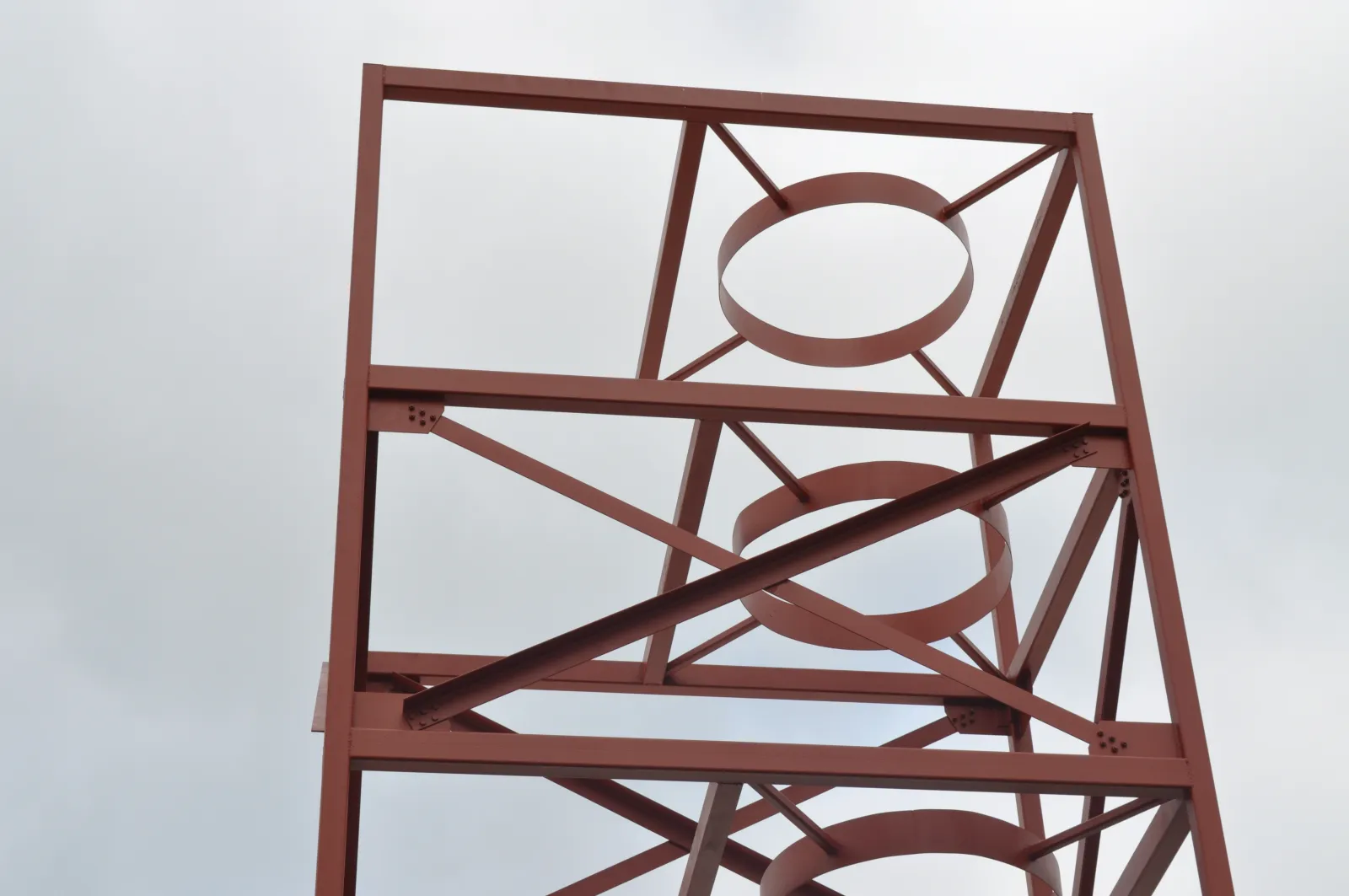 Rust-colored metal framework with circular and diagonal supports against a cloudy sky.