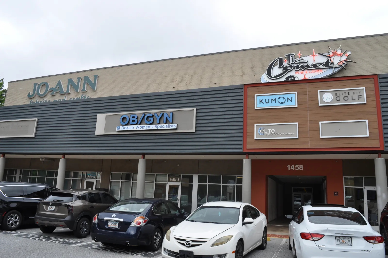 Shopping plaza exterior with storefronts including JOANN, OB/GYN, Kumon, and parking lot with cars in front.