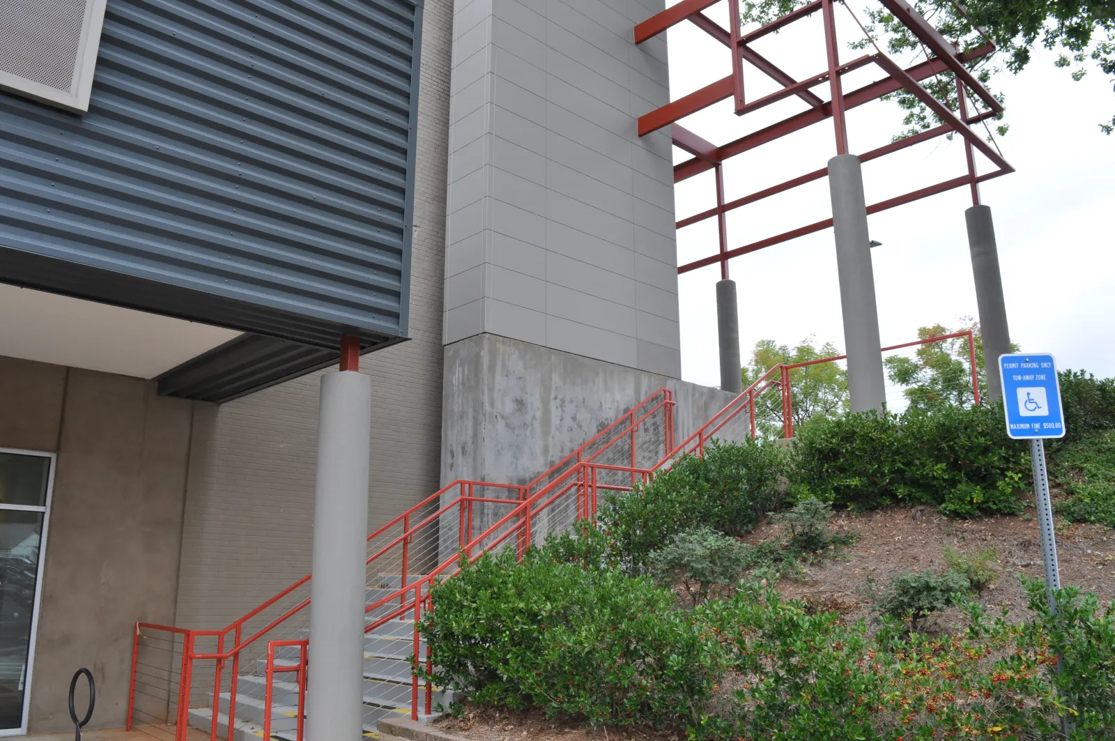 Outdoor staircase with red railings next to modern building and green shrubbery under metal canopy.