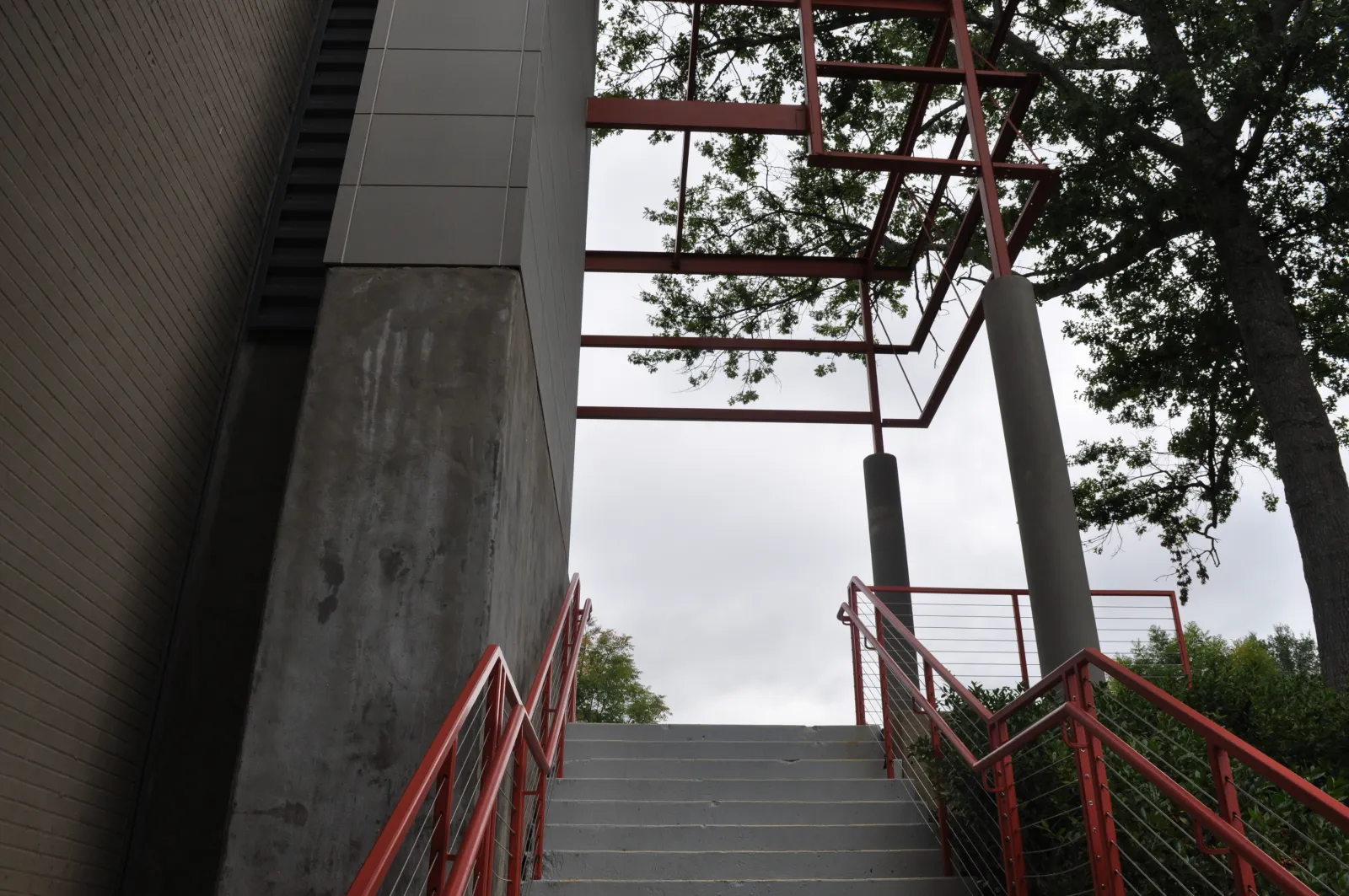 Concrete stairs with red railings lead up to a metal frame structure surrounded by trees against a cloudy sky.