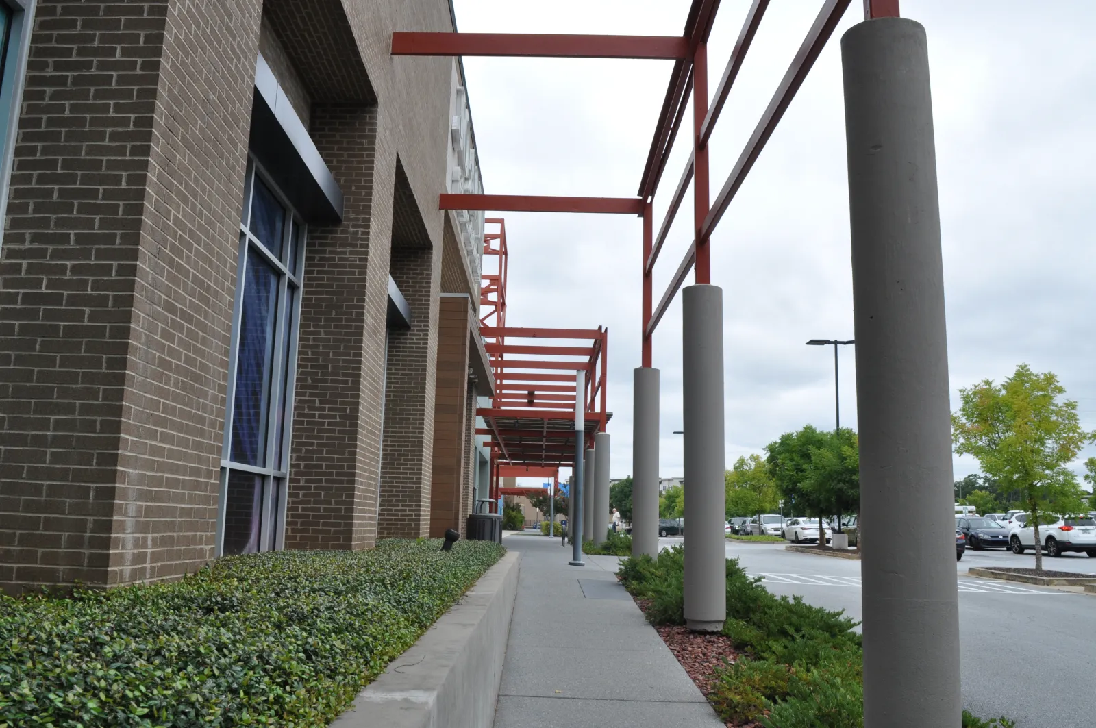 Sidewalk beside a brick building with red steel beams and concrete pillars under an overcast sky near a parking lot.