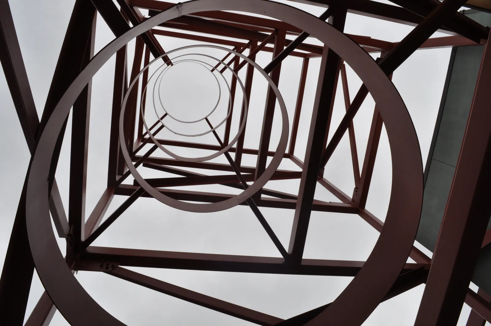 Looking upward inside a red metal tower structure with circular and diagonal beams against a cloudy sky.