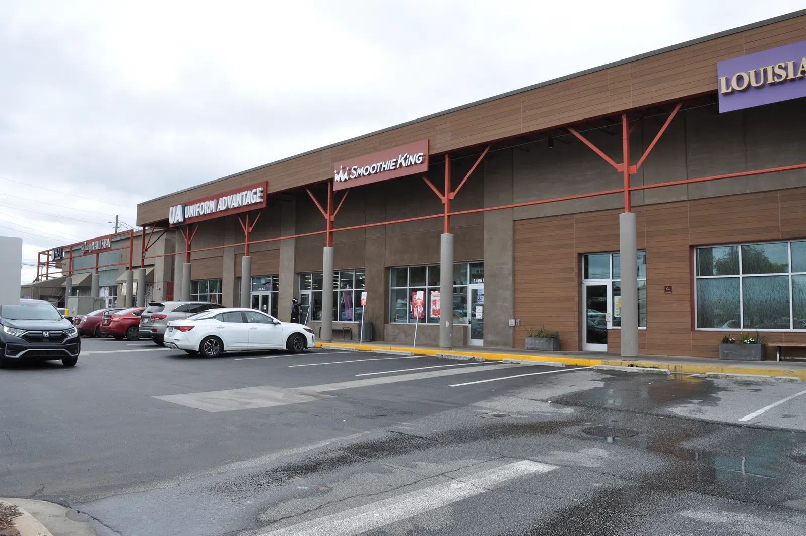 Shopping center exterior with parked cars, storefronts including Smoothie King and Uniform Advantage, cloudy sky.