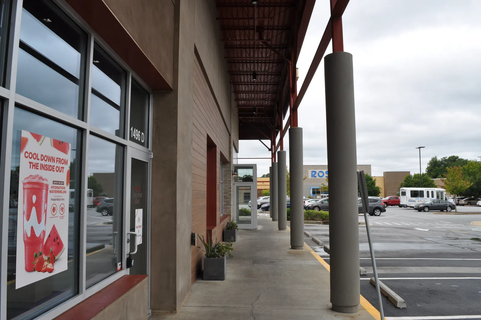 Storefront walkway with large columns, poster advertising watermelon hydration drink, and parking lot under cloudy sky.