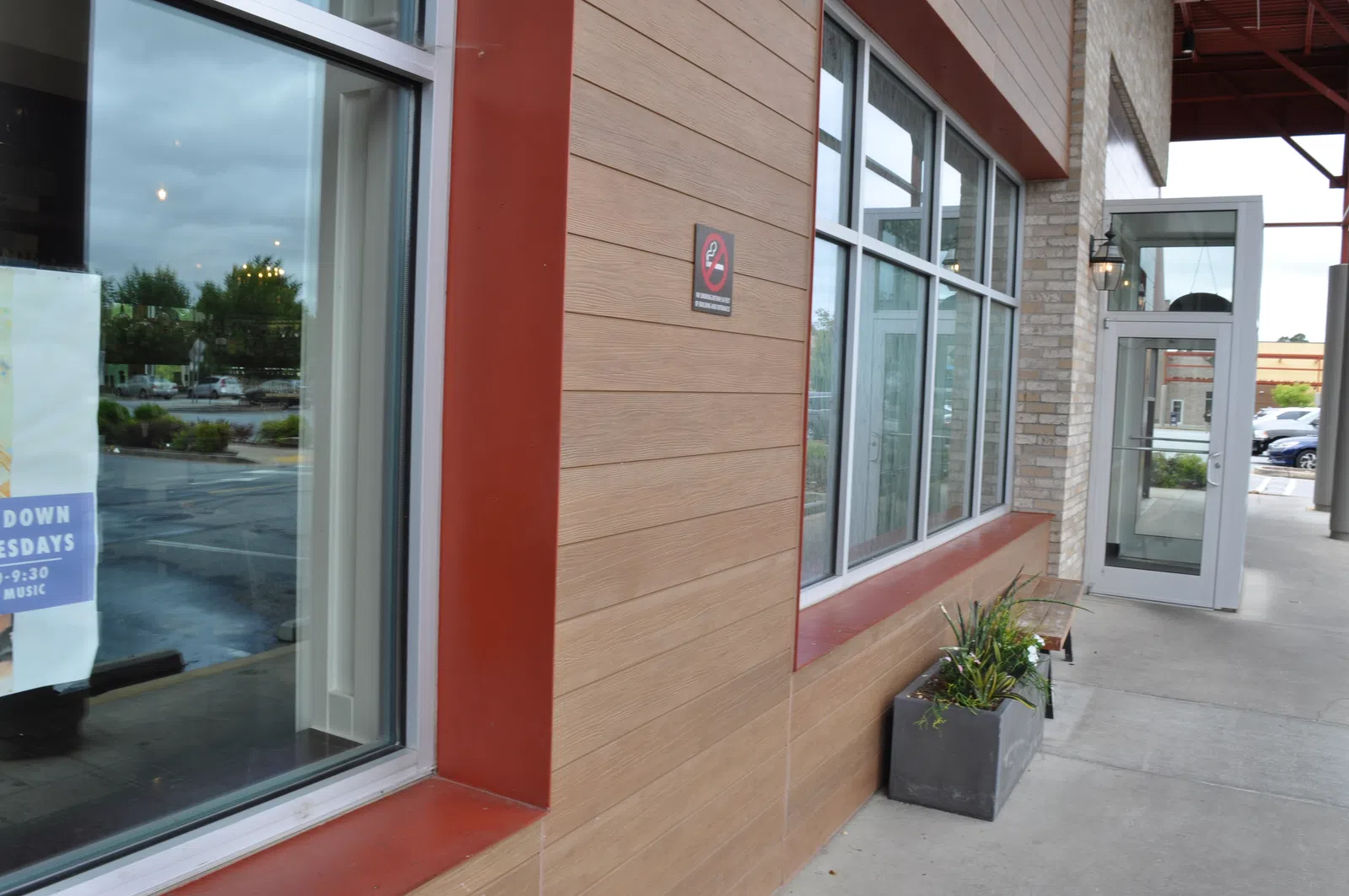 Modern restaurant exterior with large windows, tan wood paneling, potted plants, and sidewalk seating area.