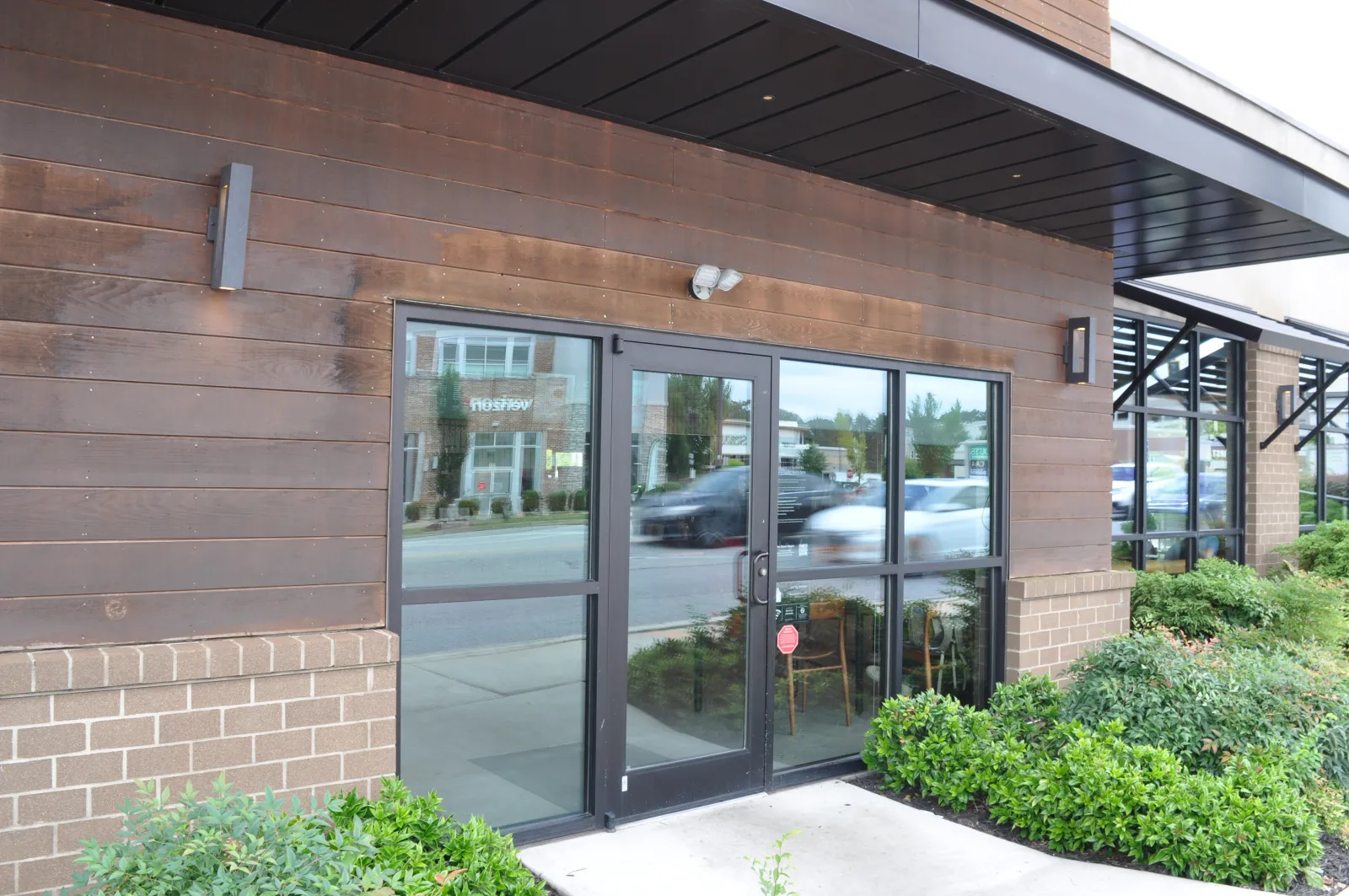 Glass double doors with black frames at storefront reflecting street and cars, surrounded by wood and brick exterior with greenery.