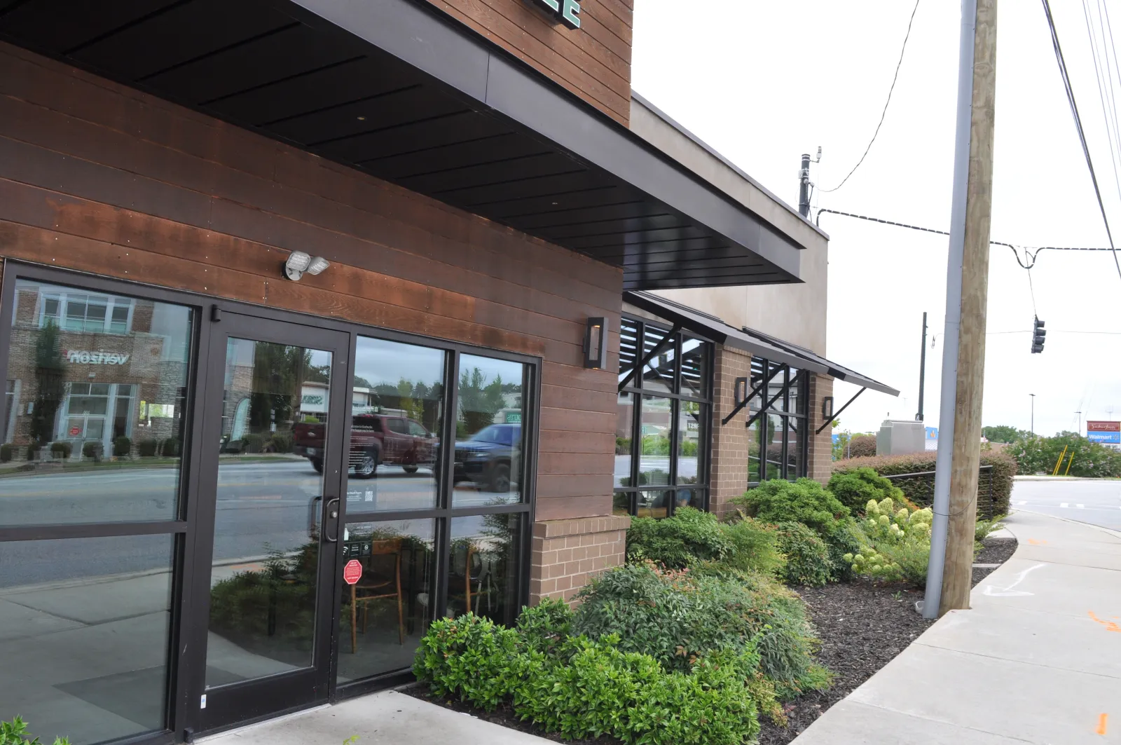 Modern brown wooden storefront with glass doors and windows, surrounded by green shrubs along a sidewalk.