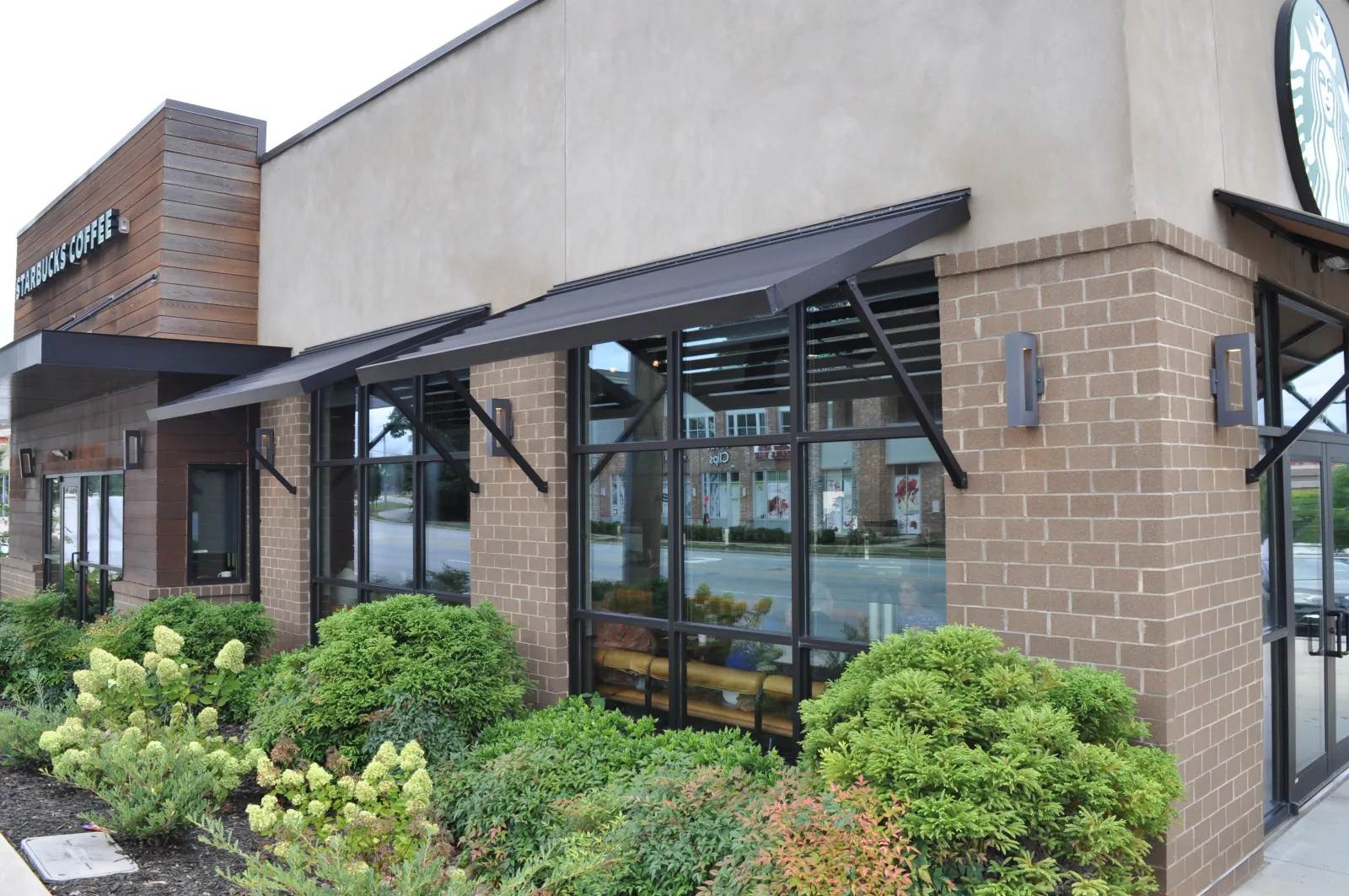 Starbucks coffee shop exterior with large windows, black awnings, brick and wood facade, and green landscaping.