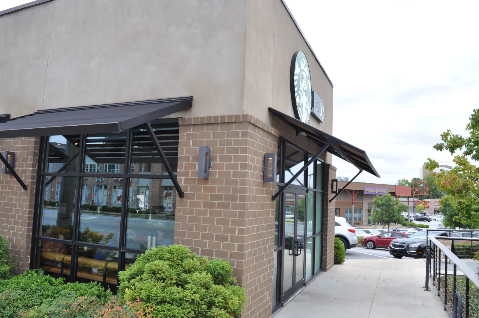 Exterior view of a Starbucks coffee shop with brick walls, large windows, awnings, and surrounding greenery on a cloudy day.
