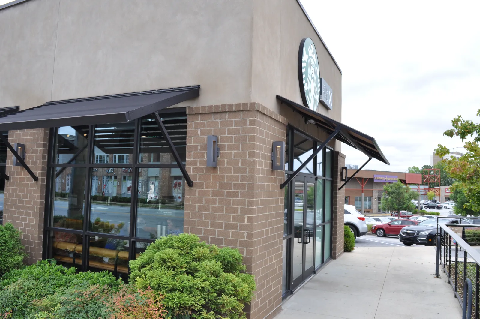 Exterior view of a Starbucks coffee shop with brick walls, large windows, and outdoor greenery near a parking lot.