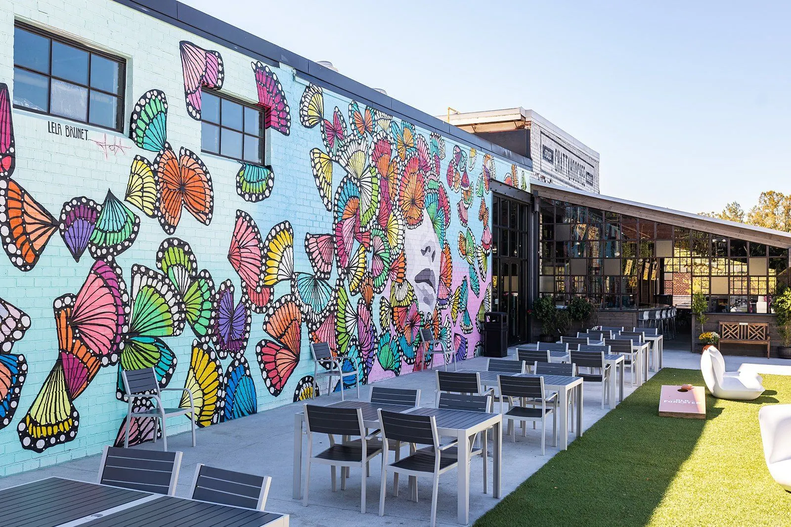 Outdoor patio with modern tables and chairs next to a colorful butterfly mural on a brick wall under clear sky