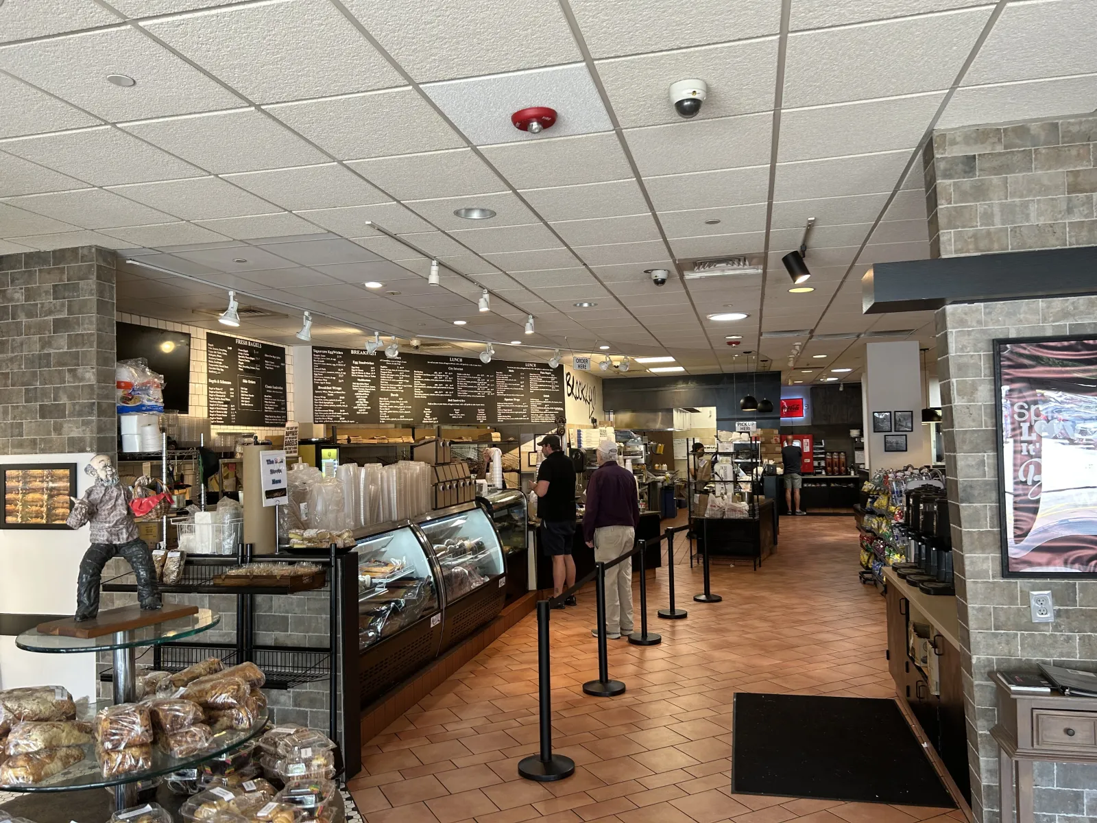 Interior of a cafe with pastry displays, customers in line, menu boards, and warm tile flooring under ceiling lights