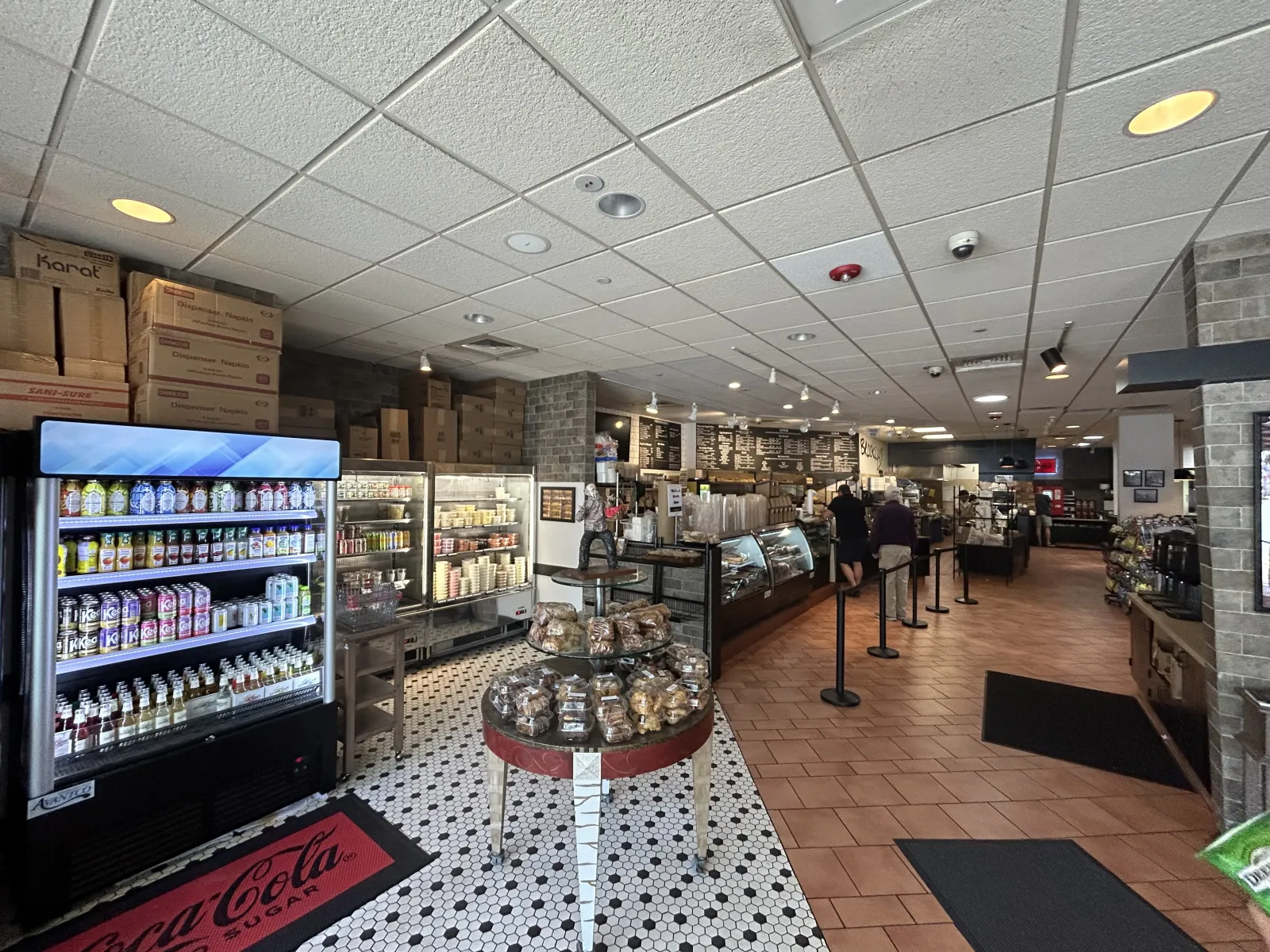 Interior of a bakery cafe with display cases, beverage fridge, baked goods table, and customers ordering at counter.
