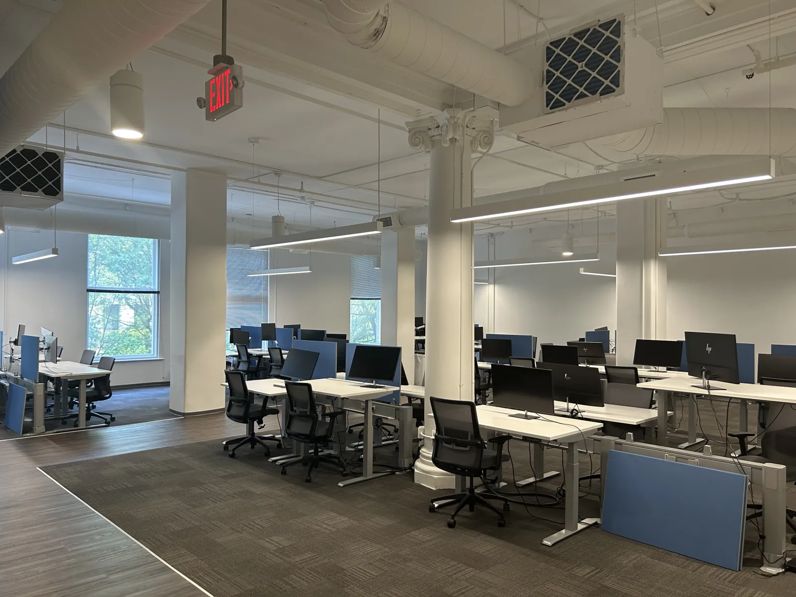 Modern office space with multiple desks, black chairs, and computer monitors under bright lighting and large windows.