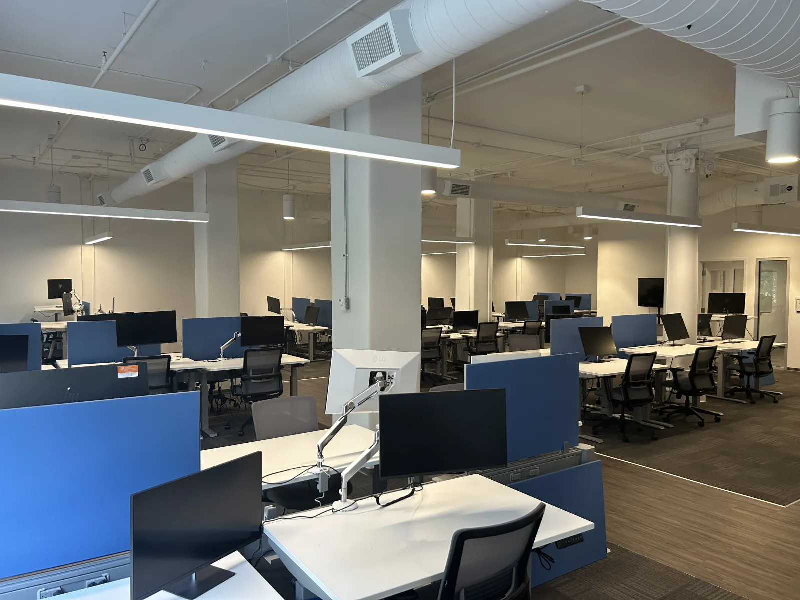 Modern open office space with rows of desks, computer monitors, ergonomic chairs, and bright overhead lighting.