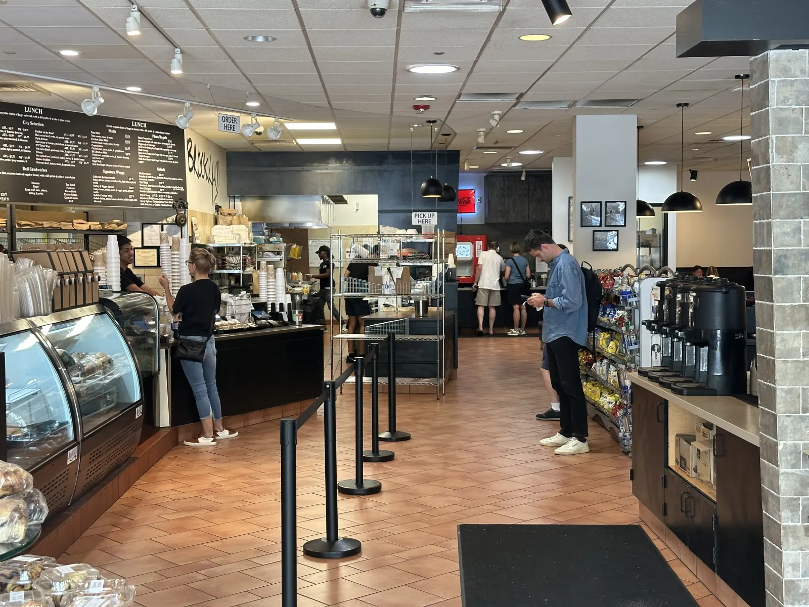 Spacious cafe interior with customers ordering, menu boards, snack shelves, and coffee station with tiled floor.
